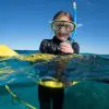A smiling child snorkeler floating on the ocean's surface with a yellow snorkel, looking at the camera.