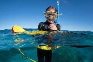 A smiling child snorkeler floating on the ocean's surface with a yellow snorkel, looking at the camera.