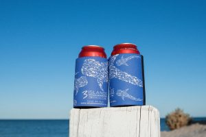 Two insulated drink cans with "3 Islands Whale Shark Dive" branding are placed on a wooden post with a beach and clear blue sky in the background.
