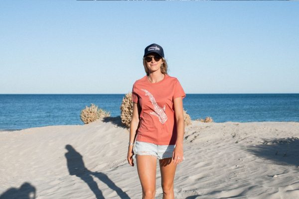 A woman stands smiling on a beach, wearing sunglasses, a cap, and a salmon-colored t-shirt with a bird design, with the ocean and clear sky in the background.