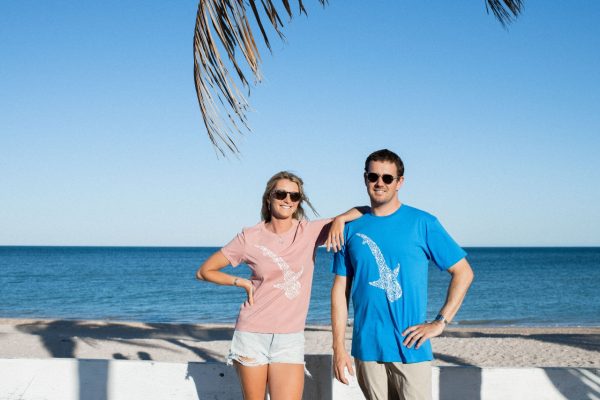 A man and a woman stand smiling on a beach with a clear sky in the background, both wearing sunglasses and t-shirts with bird designs.