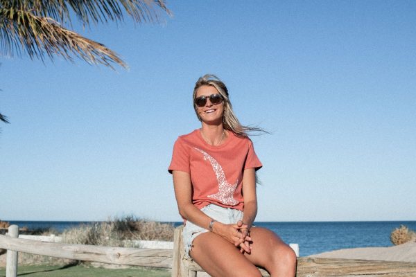 The same woman is seated on a wooden bench by the beach, wearing the coral red whale shark print t-shirt.