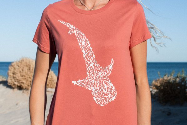 A woman in a coral red t-shirt with a white whale shark print is standing on a beach.
