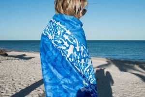 A person stands on a beach, facing away from the camera, draped in a blue towel with a white whale shark pattern, with the sea in the background.