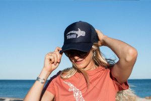 A woman adjusts her navy cap, which has a white shark design and the "3 Islands Whale Shark Dive" logo, while wearing sunglasses and a t-shirt with a bird design.