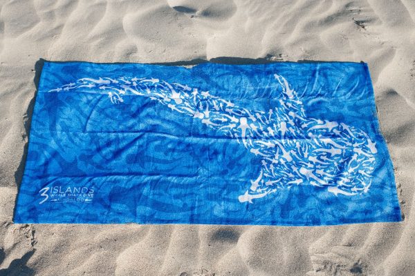 A blue towel with a whale shark pattern laid out on sandy beach, with clear visibility of the pattern and the branding of a whale shark dive company.