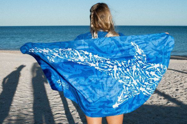 A person stands with their back to the camera, holding up a large blue towel with a whale shark pattern against a beach backdrop.
