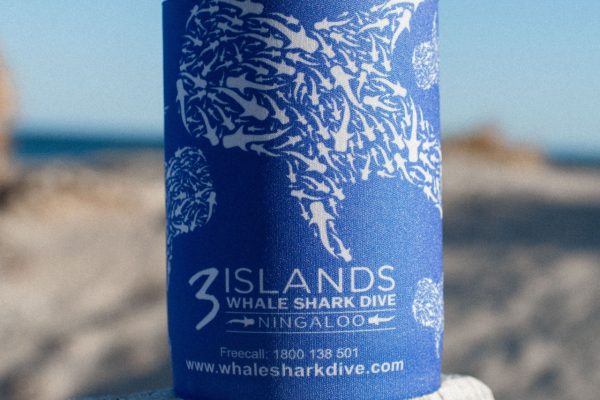 A close-up of a blue can cooler with white shark design and the "3 Islands Whale Shark Dive" branding against a blurred beach backdrop.