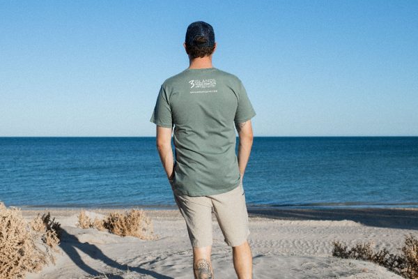 A man is seen from behind, wearing a green t-shirt with the "3 Islands Whale Shark Dive" logo and website on it, standing on a beach with the ocean in the distance.