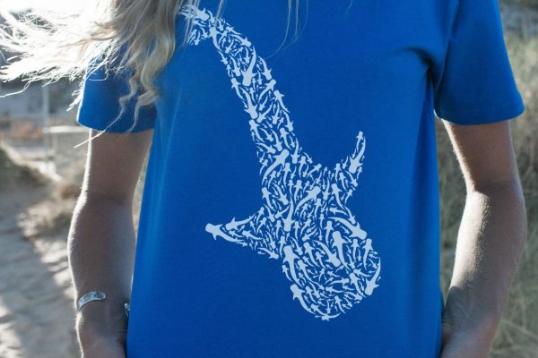 A close-up of a woman's back, showcasing a blue t-shirt with a white shark design, with a sandy beach in the background.