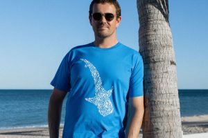 A man wearing sunglasses and a blue t-shirt with a white shark design on the front stands on a beach with the ocean and a clear sky in the background.