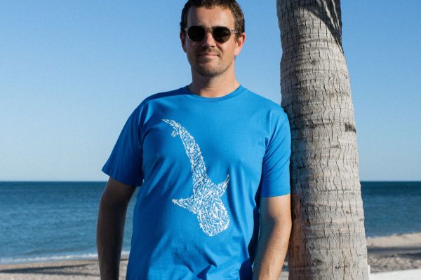 A man wearing sunglasses and a blue t-shirt with a white shark design on the front stands on a beach with the ocean and a clear sky in the background.