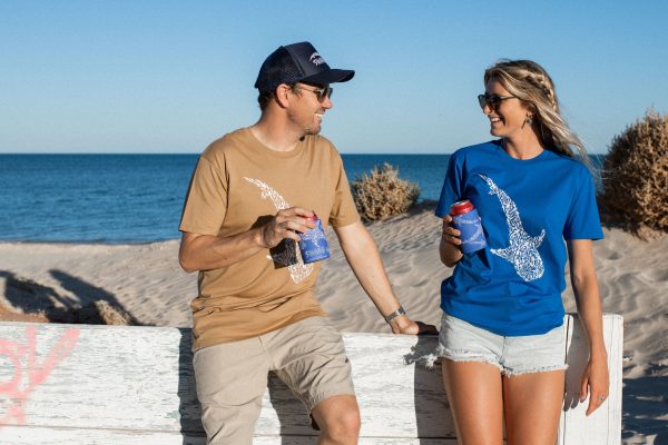 A man and woman are casually talking on a beach, both holding drinks with branded can holders, the man in a brown shirt and the woman in a blue shirt with a whale shark design.