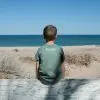 A child in a blue T-shirt with "Three Islands Whale Shark Dive Ningaloo" printed on the back, standing in front of a tree.