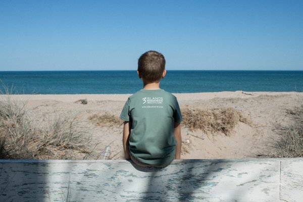 A child in a blue T-shirt with "Three Islands Whale Shark Dive Ningaloo" printed on the back, standing in front of a tree.