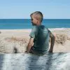 A child in a blue T-shirt with "Three Islands Whale Shark Dive Ningaloo" printed on the back, standing in front of a tree.