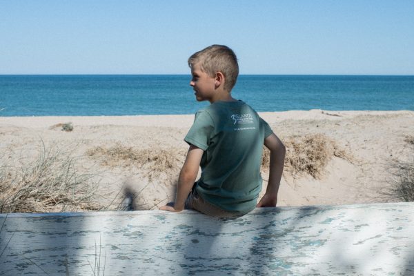 A child in a blue T-shirt with "Three Islands Whale Shark Dive Ningaloo" printed on the back, standing in front of a tree.