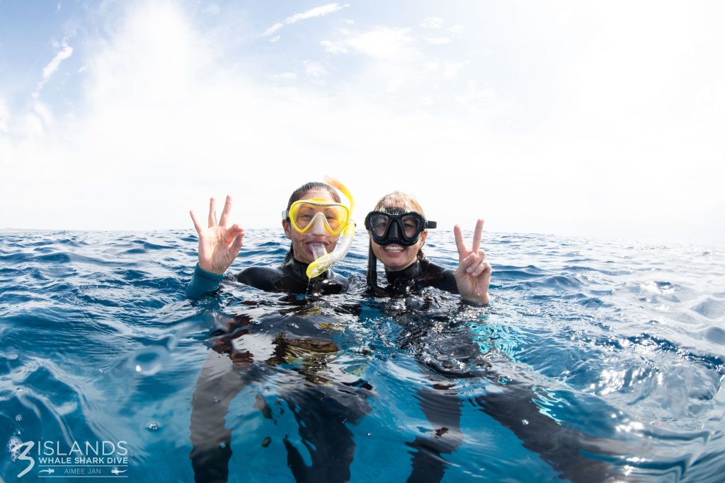 Two divers giving peace signs while floating on the ocean surface, wearing snorkeling gear.