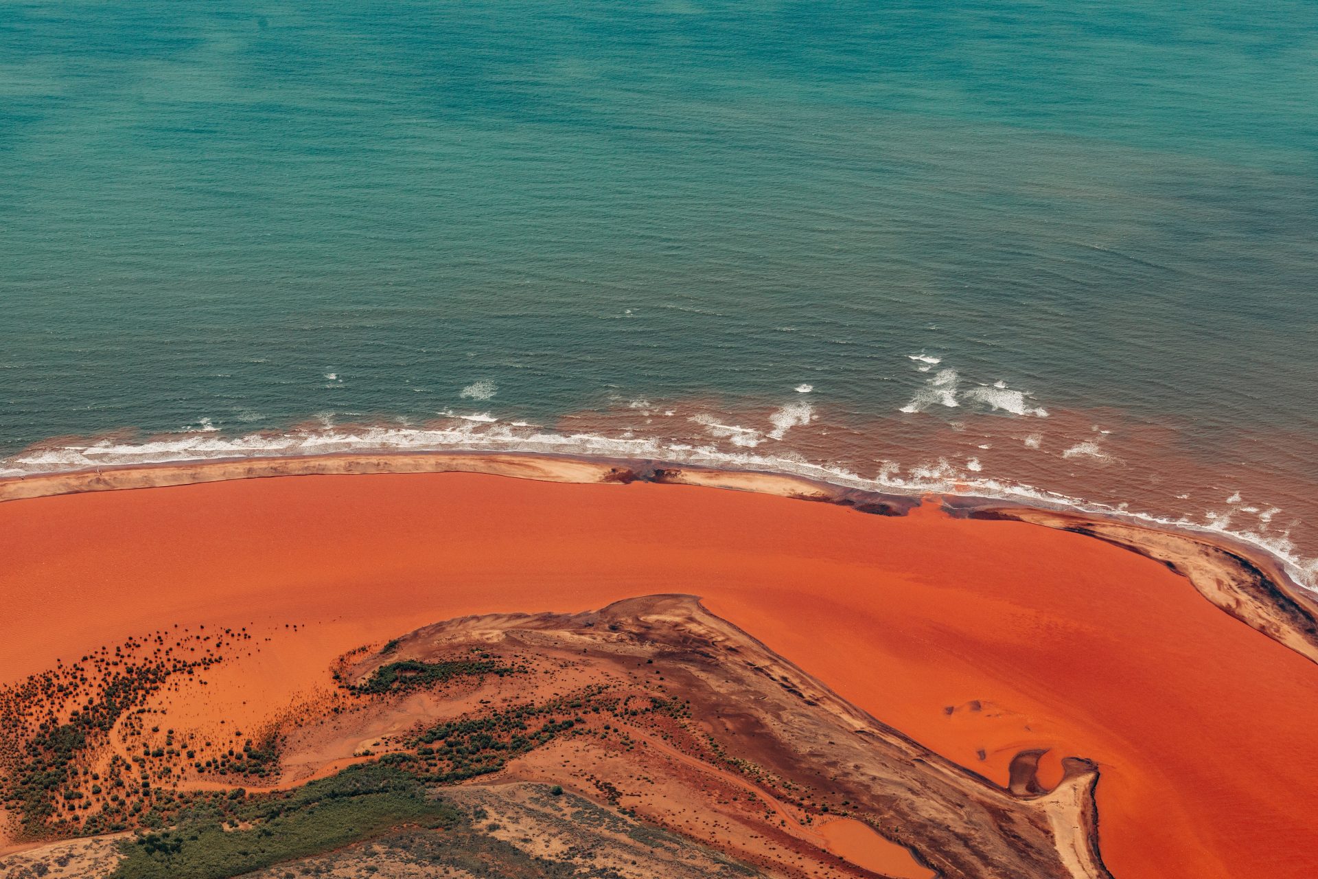 Three Island Wahle Shark Dive The Traditional Custodians of Ningaloo Aerial view of red sand beach meeting turquoise ocean, with waves and scattered green vegetation on the shoreline.