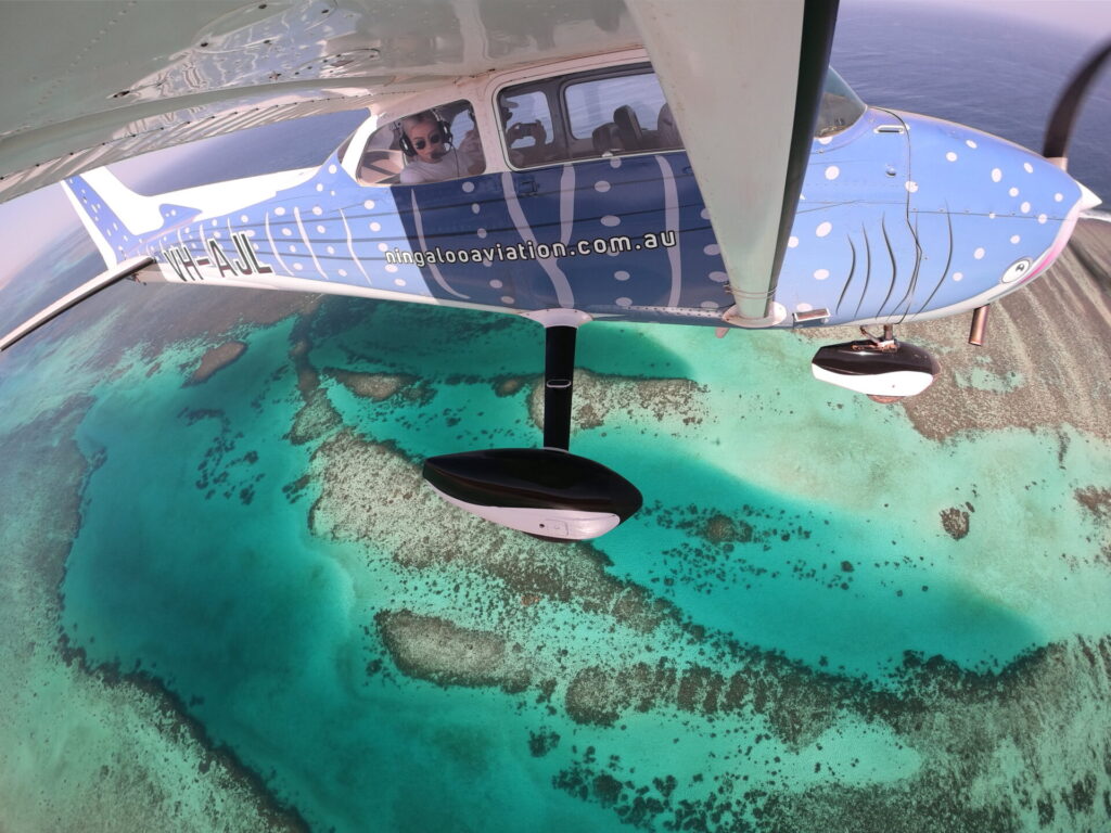 Float plane flying over Ningaloo Reef with two passengers inside, one photographing and the other looking out the window in awe.