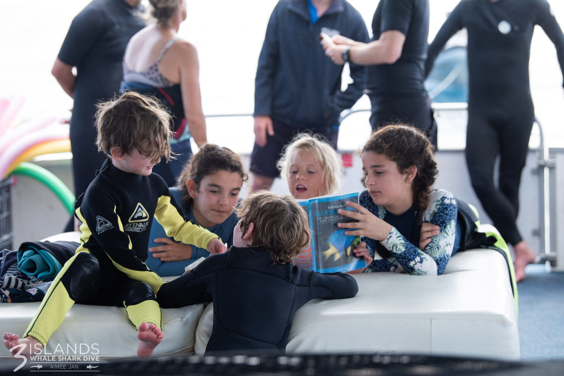 Children in wetsuits reading a marine life book aboard a whale shark tour boat, highlighting a family-friendly experience in Can Kids Go On Whale Shark Tours? What Families Need to Know.