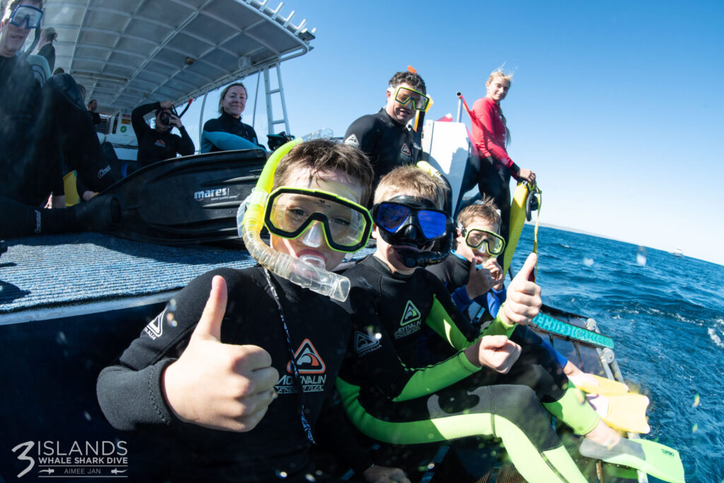Three young boys in snorkelling suits giving thumbs up on a tour boat, with a small group of snorkellers in the background.