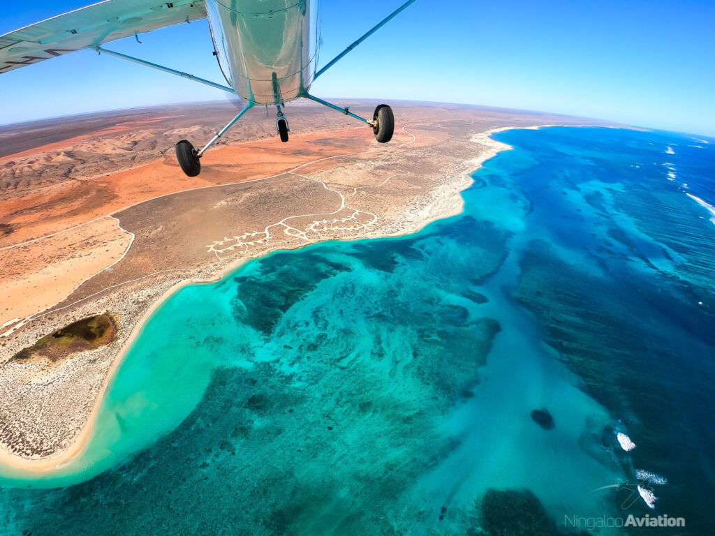 A small airplane flies over the Ningaloo Coast, showcasing the contrast between the turquoise coral reef lagoon and the red desert landscape below